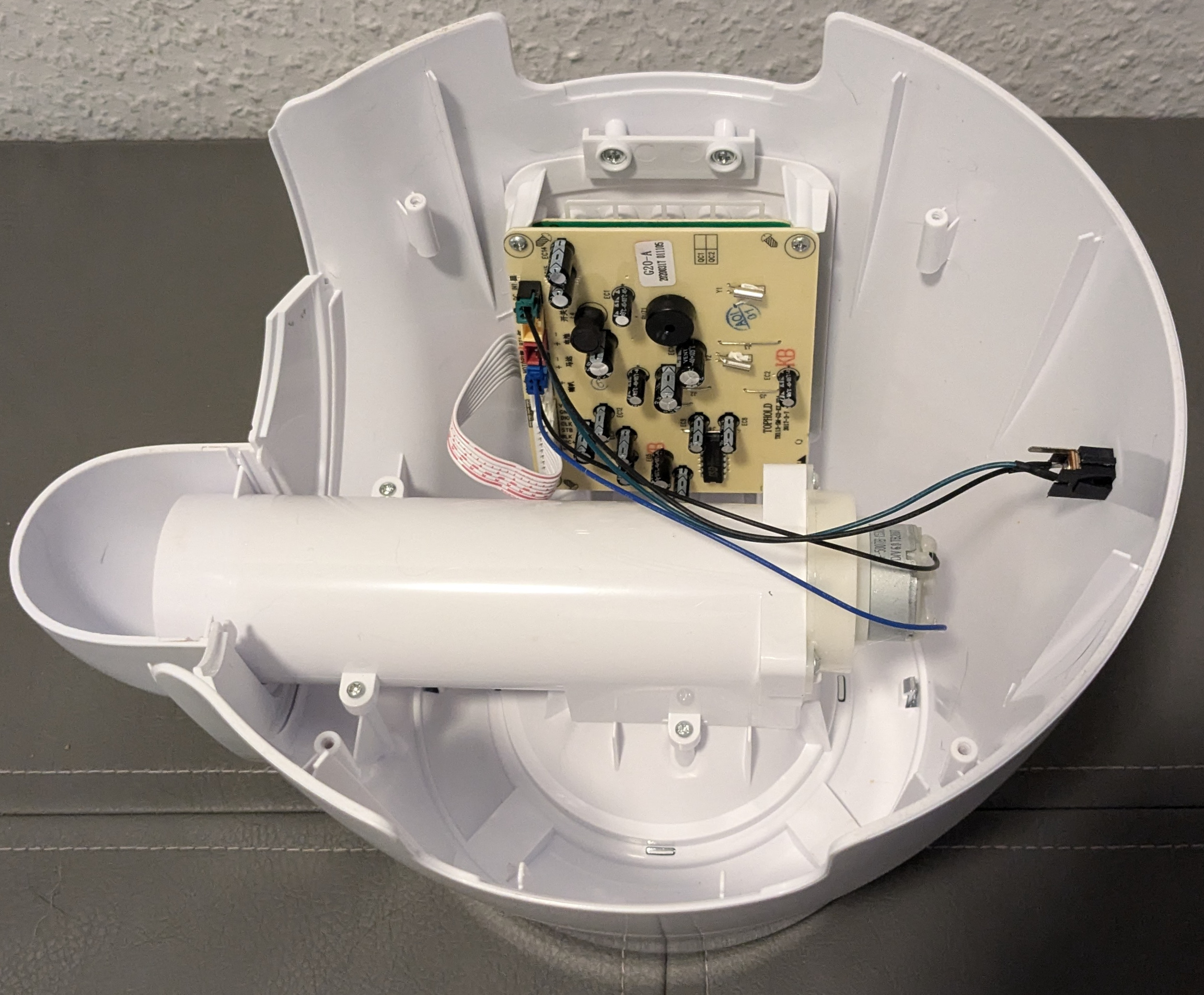 The upper half of the core, now disconnected from the bottom. The cylinder still runs down the middle. Attached to the back is a silver motor. On the far side of it, we can see a beige circuit board populated with parts. Characters in english and Chinese are visible on the board. We can also see a black plastic item attached to the outer wall of the cylinder behind the end of the cylinder on the right of the image. This is where the power cord normally plugs in.